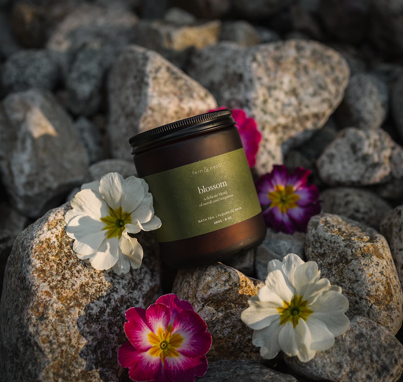 A jar of blossom bath tea laying on rocks surrounded by flowers